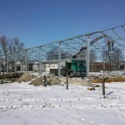 Wznoszenie szkieletu hali z metalowych profili w zimowej scenerii, widoczny dźwig z operatorem i dwaj monterzy na rusztowaniu, fundamenty i zasypany śniegiem teren budowy.