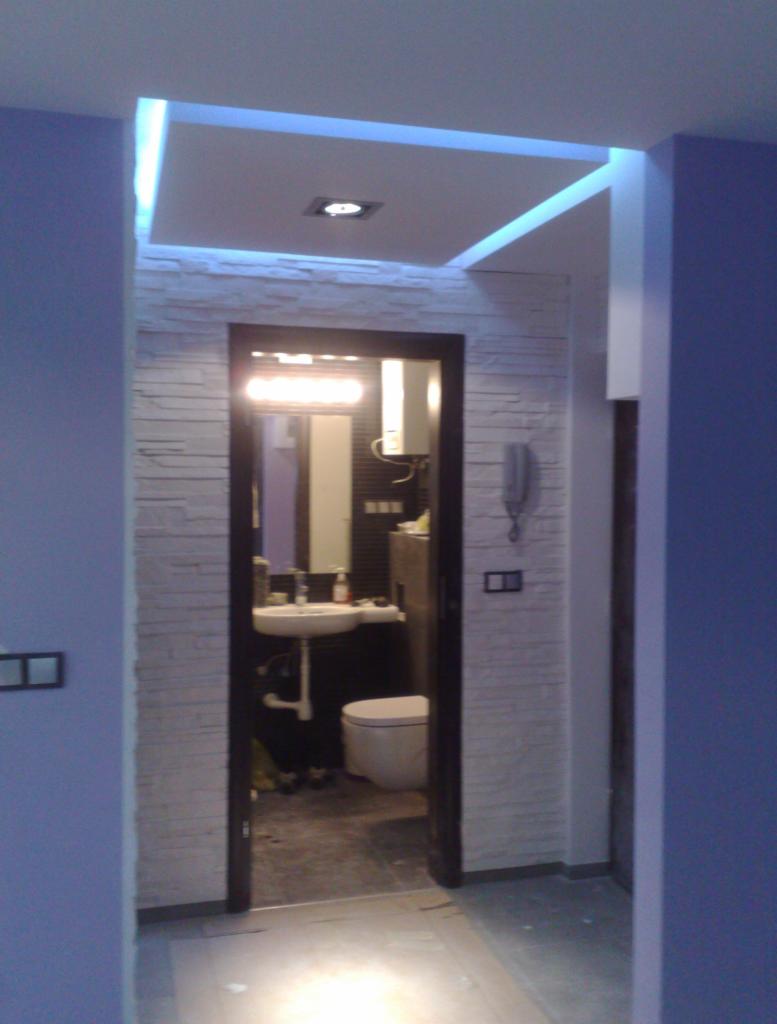 Wykończone wnętrze łazienki z podwieszanym sufitem z oświetleniem LED, ściany wyłożone dekoracyjną płytką imitującą kamień, widok przez otwarte drzwi.