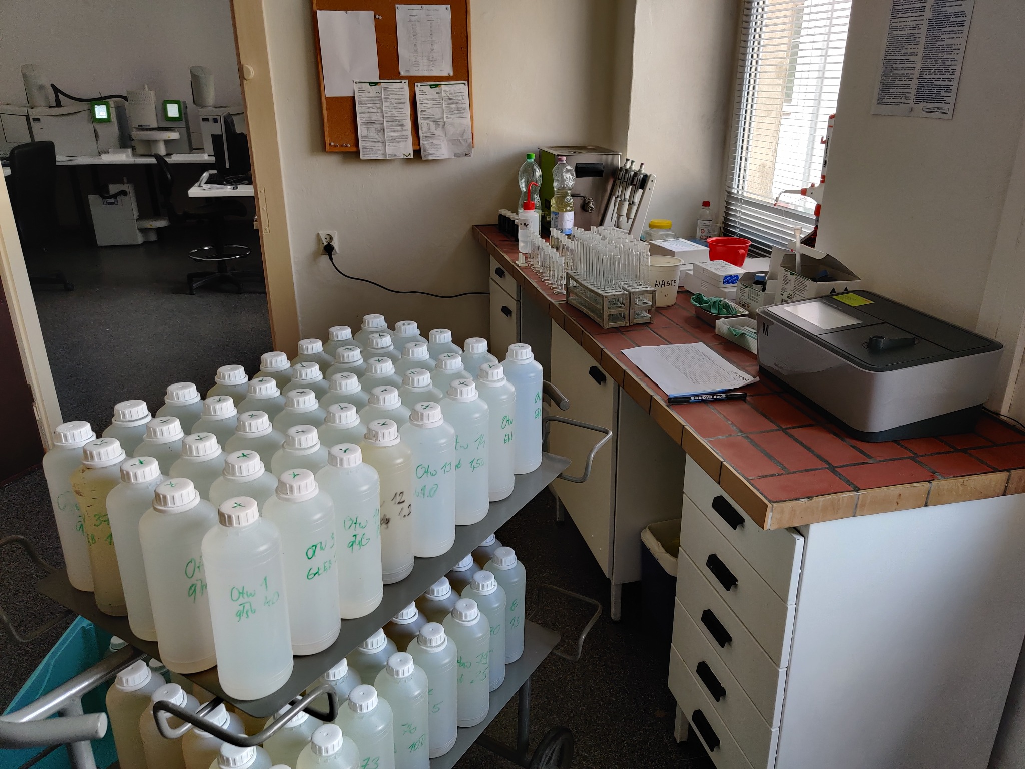 Wózek z licznymi plastikowymi butelkami z próbkami wody, opisane markerem, w laboratorium z biurkiem, probówkami i analizatorem.