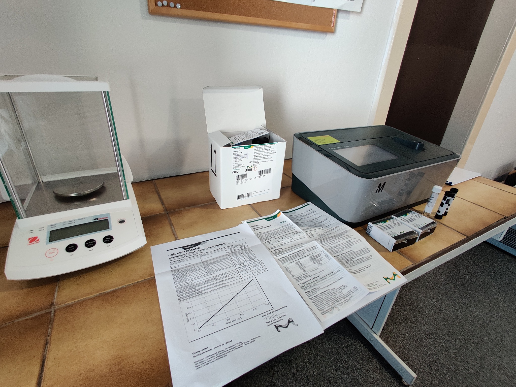 Laboratorium: waga analityczna, otwarte pudełko z odczynnikami, spektrofotometr, fiolki z odczynnikami i dokumentacja testowa z wykresem kalibracyjnym na stole.