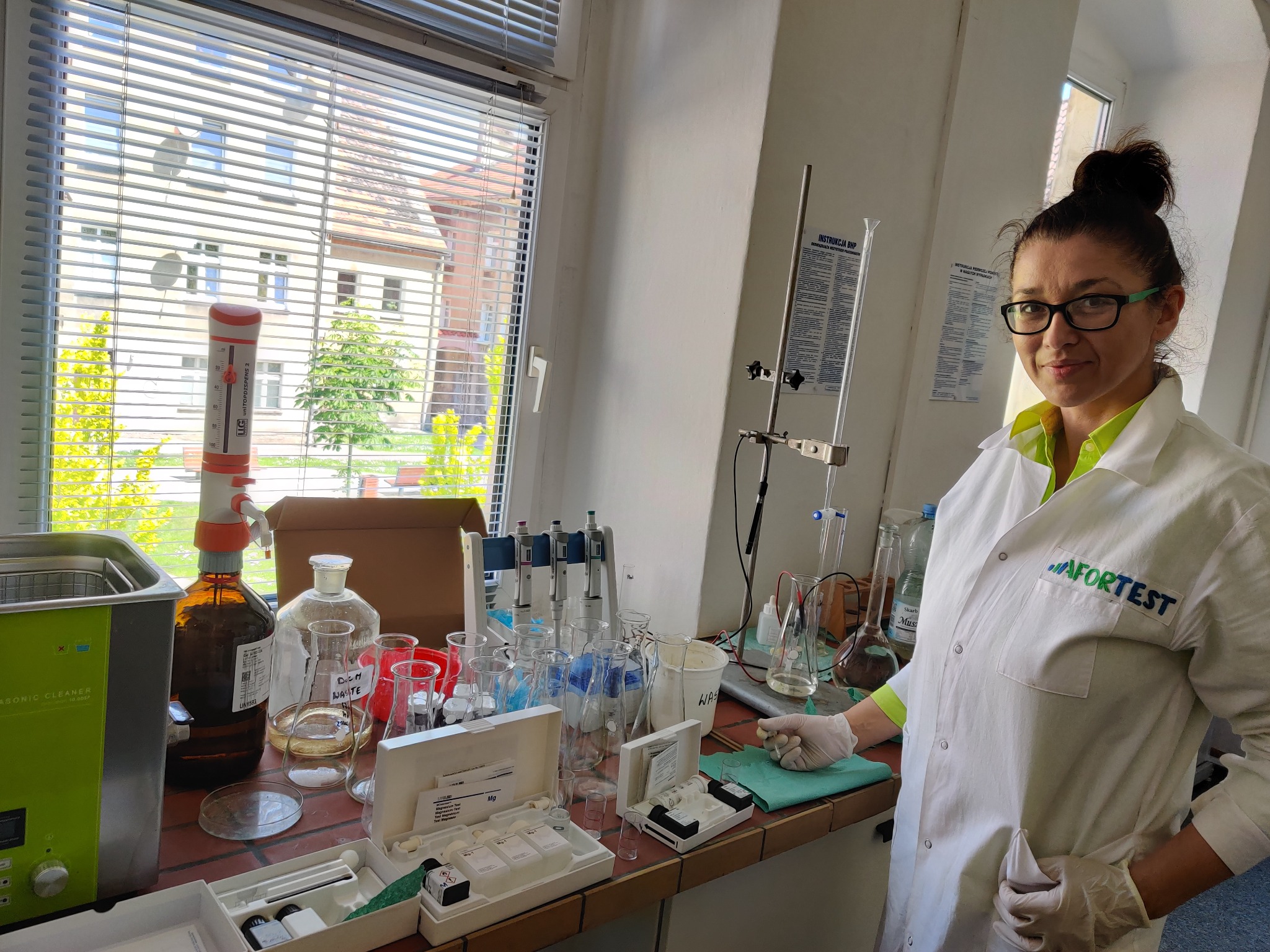 Kobieta w okularach i białym fartuchu laboratoryjnym z logo 'AFORTEST' stoi w laboratorium, otoczona szkłem laboratoryjnym, odczynnikami i aparaturą, w tle okno z widokiem na budynek.