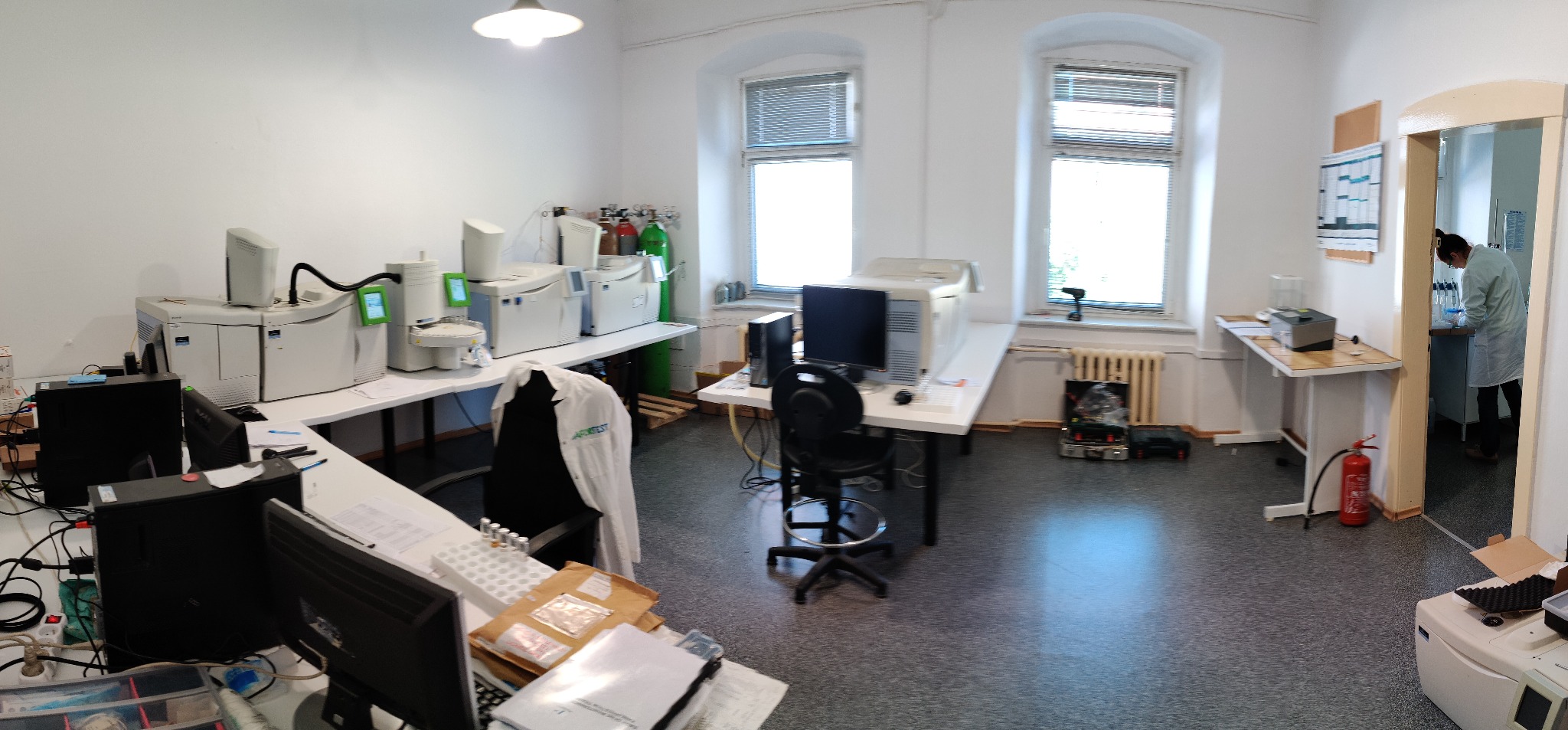 Panorama laboratorium z aparaturą analityczną, komputerami i pracownikiem w fartuchu, widoczne butle z gazami technicznymi i gaśnica.