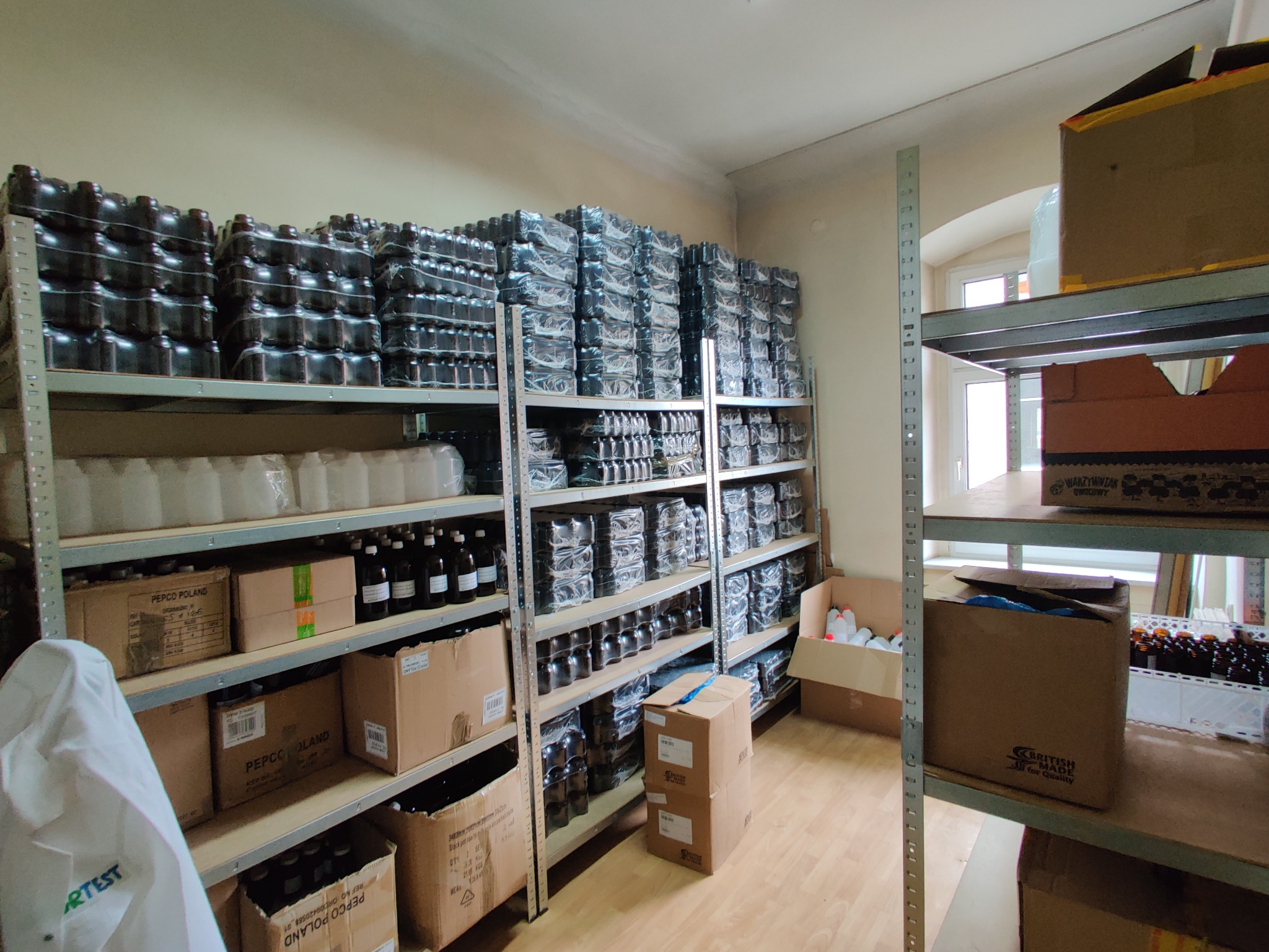 Magazyn z zapasem ciemnych butelek na metalowych regałach, częściowo zapakowanych w folię, obok kartony z napisem 'PEPCO POLAND' i 'BRITISH MADE for Quality'.