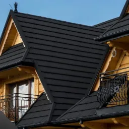 Częściowy widok drewnianego domu z ciemnym dachem z metalowych paneli, ozdobne balustrady balkonowe, błękitne niebo w tle, logo firmy w lewym dolnym rogu.