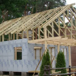 Konstrukcja domu szkieletowego w trakcie budowy w Kole, widoczne drewniane elementy nośne i ściany zabezpieczone folią budowlaną, w tle zieleń i zabudowania.