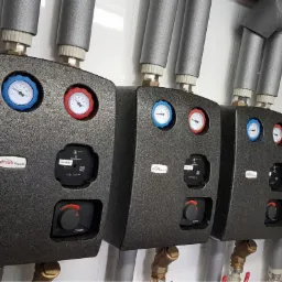 Trzy czarne moduły rozdzielacza ogrzewania podłogowego z manometrami w kolorach niebieskim i czerwonym, podłączone do rur w izolacji, zamontowane na białych płytkach.