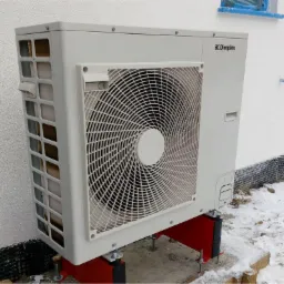 Biała pompa ciepła marki Dimplex zamontowana na czerwonych wspornikach przy ośnieżonym budynku z widocznym oknem zabezpieczonym folią.