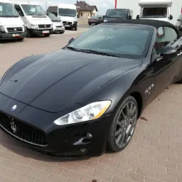 Czarny kabriolet Maserati GranCabrio z otwartym dachem, zaparkowany na brukowanym placu, z białymi vanami w tle.
