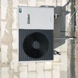 Zewnętrzna jednostka grzewcza Vaillant aroTHERM zamontowana na ścianie z białych bloczków betonowych, widoczne przewody i rury przyłączeniowe.