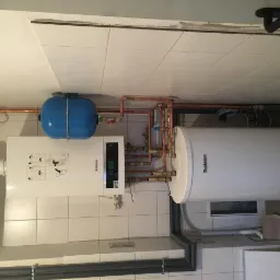 Biały kocioł grzewczy Buderus z niebieskim zbiornikiem wyrównawczym, połączony miedzianymi rurami w pomieszczeniu z białymi kafelkami.