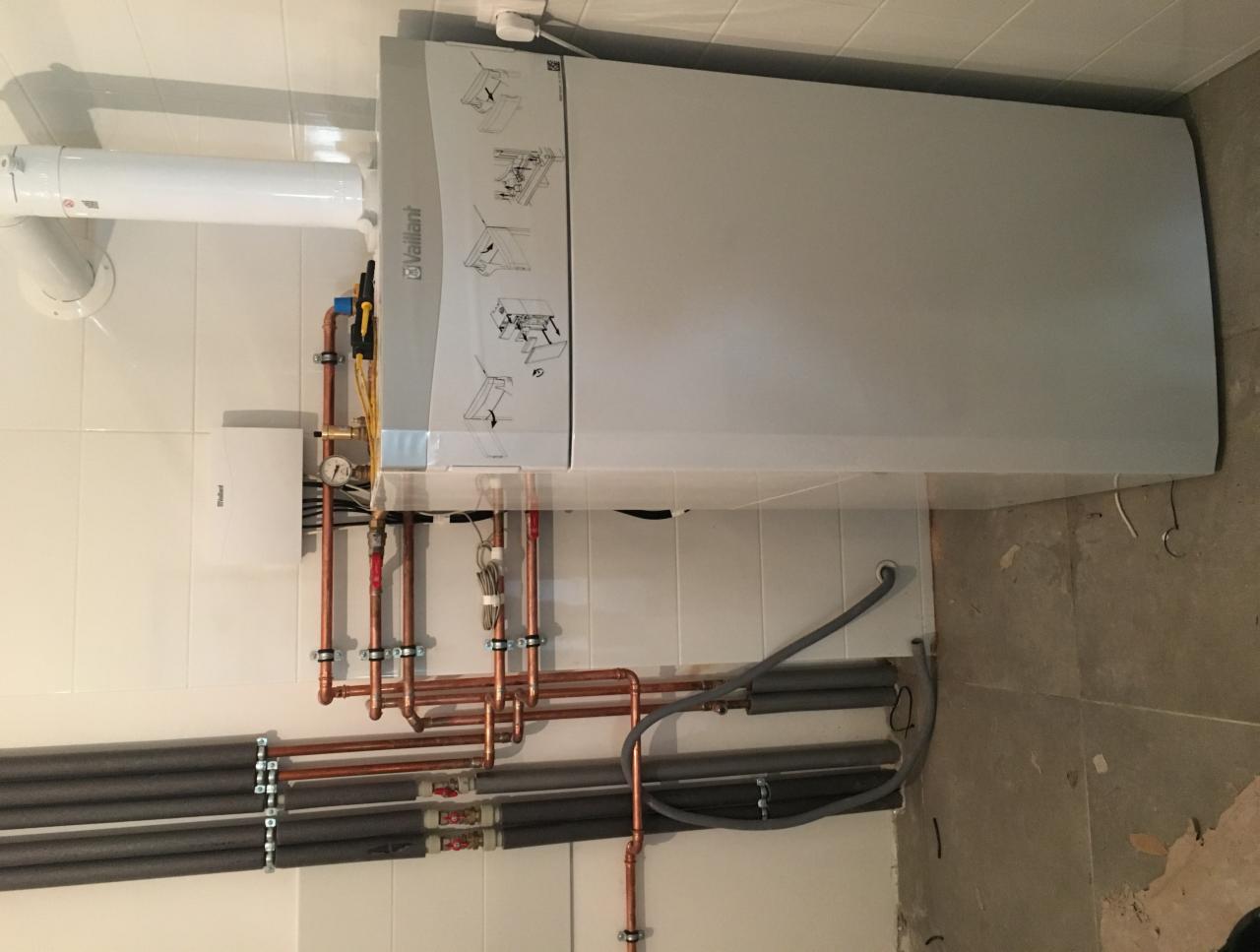 Biały wiszący kocioł gazowy marki Vaillant z podłączonymi miedzianymi rurami i szarymi rurami izolowanymi, zamontowany na ścianie z białych płytek, widoczne elementy instalacji.