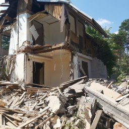 EFEKT Iwona Kamińska - Zrujnowany, dwukondygnacyjny dom z ceglanym kominem, otoczony gruzem i zielenią. Widoczne fragmenty ścian, okien i dachu po rozbiórce. Błękitne niebo w tle.