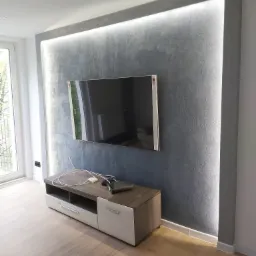 Szara ściana dekoracyjna z podświetleniem LED i zawieszonym telewizorem. Pod ścianą szafka RTV w odcieniach bieli i drewna. Parkiet na podłodze. Nowoczesne wykończenie wnętrza.