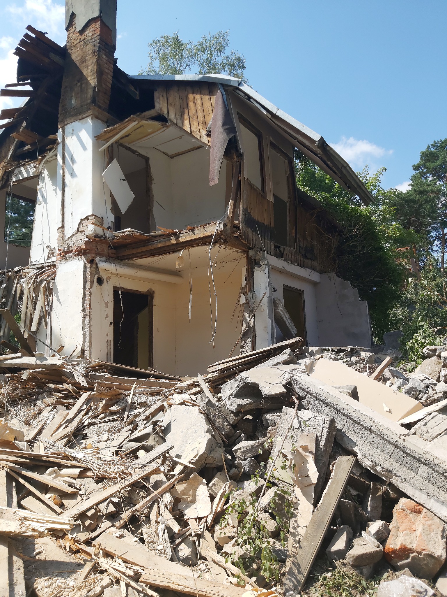 Zrujnowany, dwukondygnacyjny dom z ceglanym kominem, otoczony gruzem i zielenią. Widoczne fragmenty ścian, okien i dachu po rozbiórce. Błękitne niebo w tle.