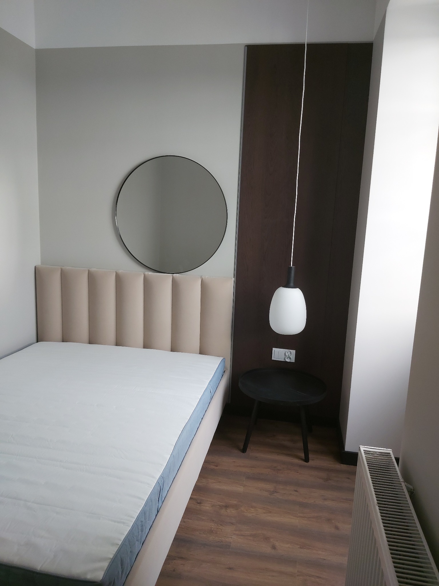 Nowoczesna sypialnia: łóżko z tapicerowanym zagłówkiem, okrągłe lustro, lampa wisząca nad stolikiem nocnym, grzejnik. Drewniana podłoga i panel ścienny.