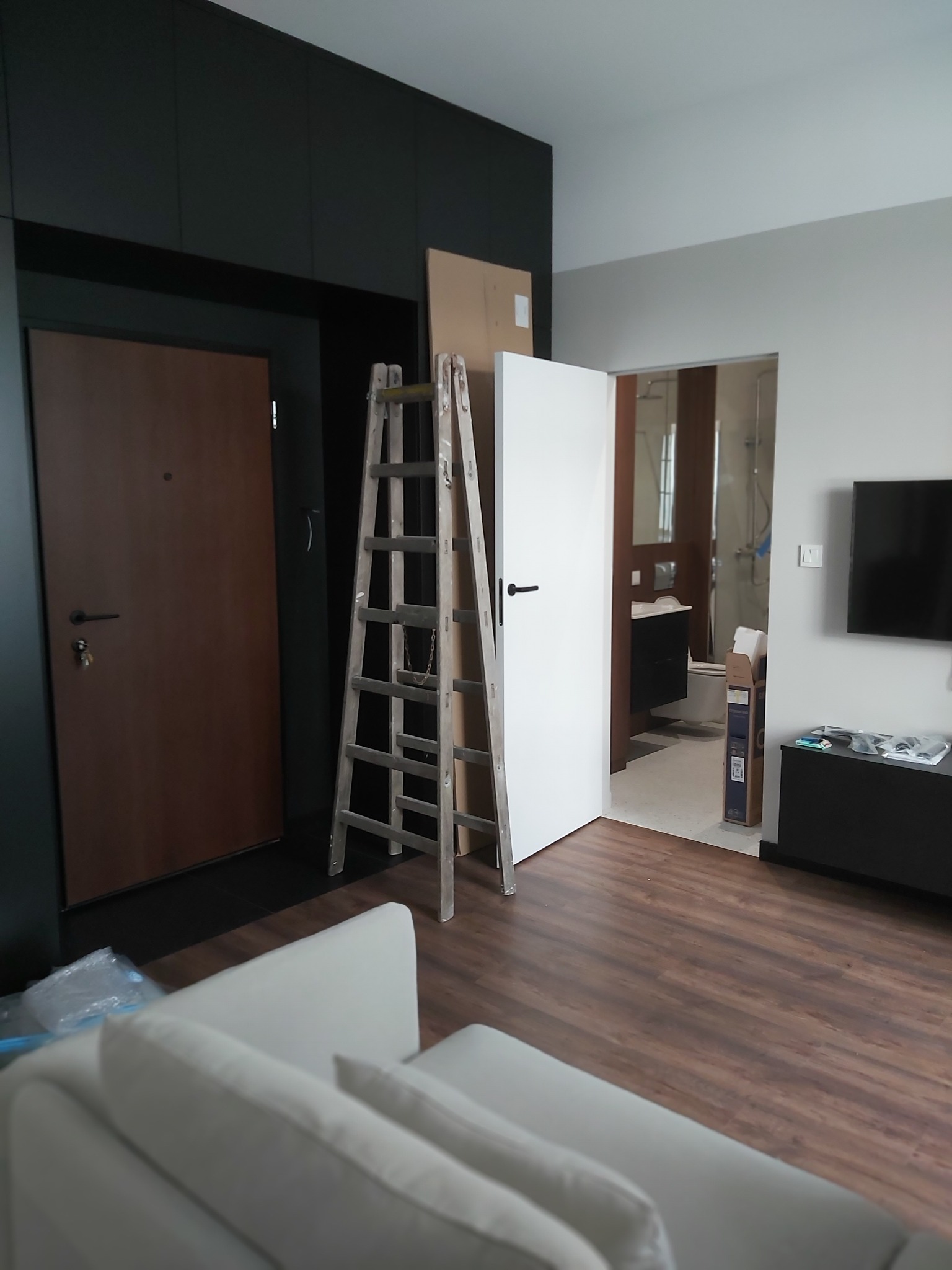 Wnętrze w trakcie wykończenia: drewniane drzwi, białe drzwi wewnętrzne, drabina malarska. W tle widoczna łazienka i częściowo sofa. Parkiet na podłodze.