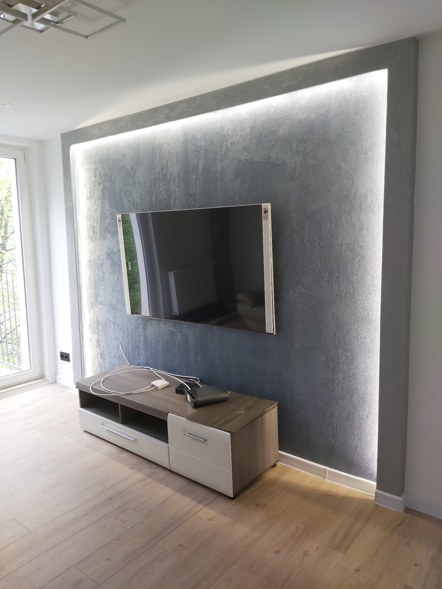 Szara ściana dekoracyjna z podświetleniem LED i zawieszonym telewizorem. Pod ścianą szafka RTV w odcieniach bieli i drewna. Parkiet na podłodze. Nowoczesne wykończenie wnętrza.