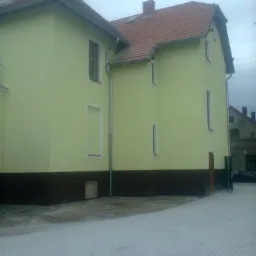 Żółty budynek po nałożeniu tynku, z brązowym pasem w dolnej części elewacji i czerwoną dachówką, widoczny z perspektywy chodnika wyłożonego szarą kostką.