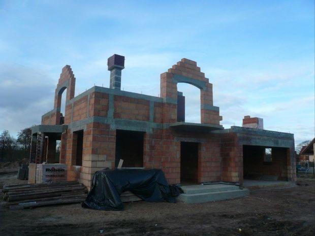Surowy stan domu z czerwonej cegły, z widocznymi otworami okiennymi i kominem w budowie, częściowo zakryty czarną folią, z paletą cegieł Roben i drewnianymi belkami na pierwszym planie.