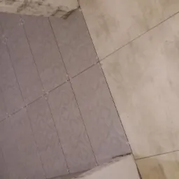 Świeżo ułożone, szare płytki na ścianie z krzyżykami dystansowymi, kontrastujące z beżowymi płytkami na podłodze, w surowym wnętrzu łazienki w trakcie remontu.