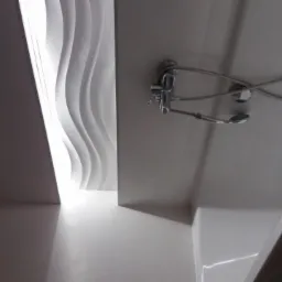 Nowoczesna łazienka z dekoracyjną ścianą o falistym wzorze i zamontowaną armaturą prysznicową. Minimalistyczny design, jasne kolory, nietypowe ujęcie.