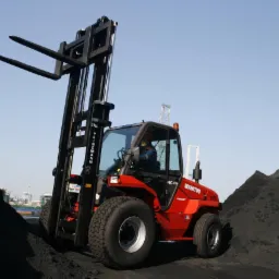 Czerwony wózek widłowy Manitou MC 40 z operatorem w kabinie, stojący na hałdzie czarnego materiału sypkiego, z widocznymi w tle dźwigami portowymi.