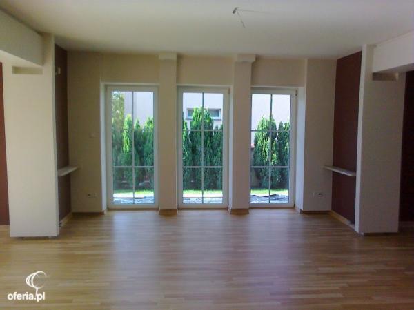 Pusty pokój z parkietem, trzema oknami z widokiem na zielony ogród i częściowo pomalowanymi ścianami w kolorach beżowym i brązowym.