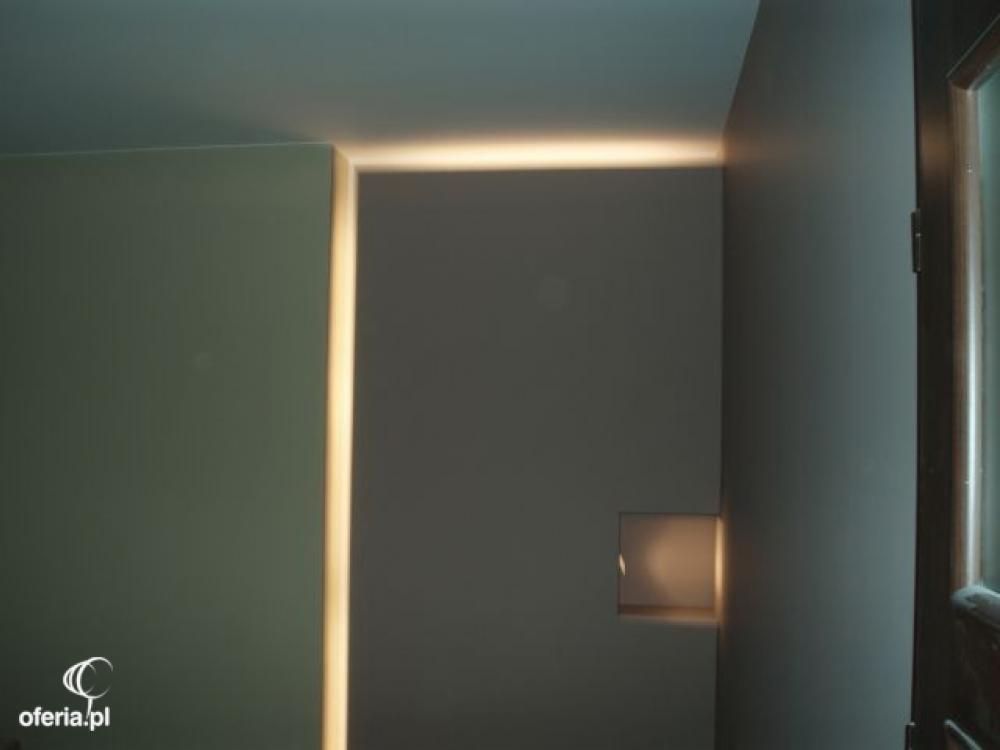 Wnęka oświetleniowa w szarej ścianie, widok z boku, minimalistyczne wykończenie, widoczny fragment stolarki okiennej.