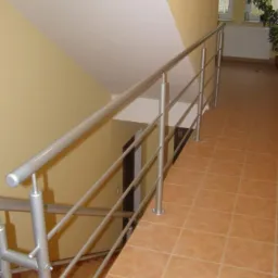 Wewnętrzna balustrada schodowa ze stali nierdzewnej, z widocznymi mocowaniami do podłogi z płytek ceramicznych w kolorze terakoty, ściany w odcieniach beżu i kremu, widok z góry na dół.