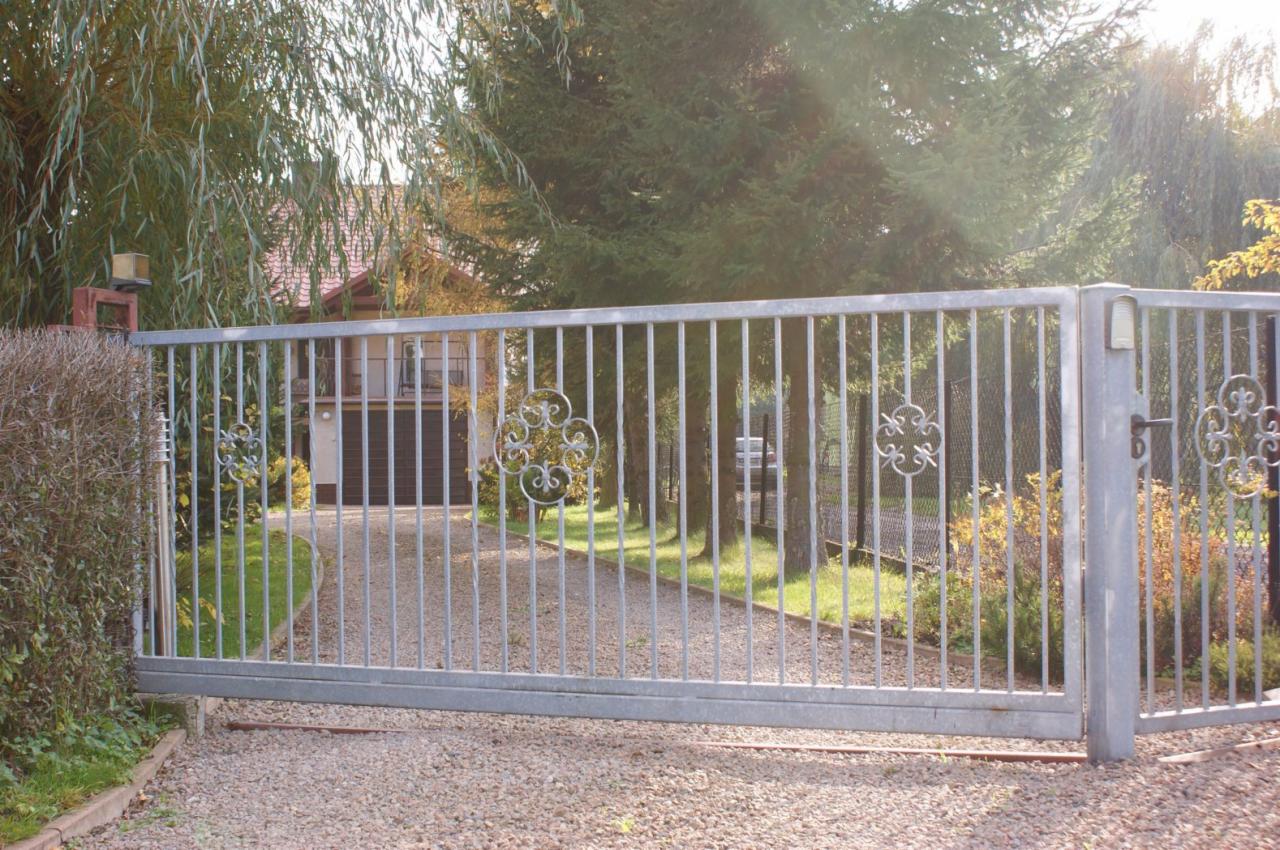 Szara, metalowa brama wjazdowa z ozdobnymi elementami na tle domu jednorodzinnego w Kłaju. Brama z pionowych prętów, widoczny podjazd wysypany żwirem.