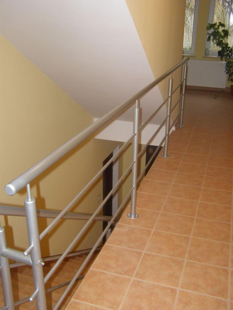 Wewnętrzna balustrada schodowa ze stali nierdzewnej, z widocznymi mocowaniami do podłogi z płytek ceramicznych w kolorze terakoty, ściany w odcieniach beżu i kremu, widok z góry na dół.