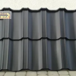 Czarna dachówka modułowa marki de Lux Husaria, ułożona na brukowanej powierzchni, z naklejką producenta w lewym górnym rogu, na tle jasnej ściany.