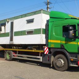 P.H.U. JANTRUCK s.c.Sebastian i Marcin Jankowiak - Zielona laweta Scania z napisem 'Pomoc Drogowa' przewozi biało-zielony domek mobilny na niskopodwoziowej platformie. Widok z boku, słoneczny dzień.