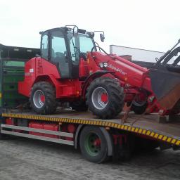 P.H.U. JANTRUCK s.c.Sebastian i Marcin Jankowiak - Zielona ciężarówka z czerwoną ładowarką teleskopową Schäffer 9510T na platformie. Ładunek zabezpieczony łańcuchami, widoczne detale opon i konstrukcji pojazdów.