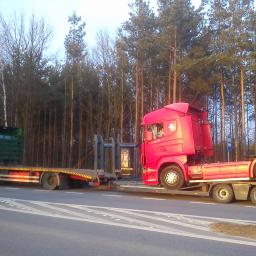 P.H.U. JANTRUCK s.c.Sebastian i Marcin Jankowiak - Dwa samochody ciężarowe typu laweta: zielony i czerwony, jadące obok siebie po drodze. W tle wysokie drzewa. Czerwony pojazd przewozi kabinę ciężarówki.