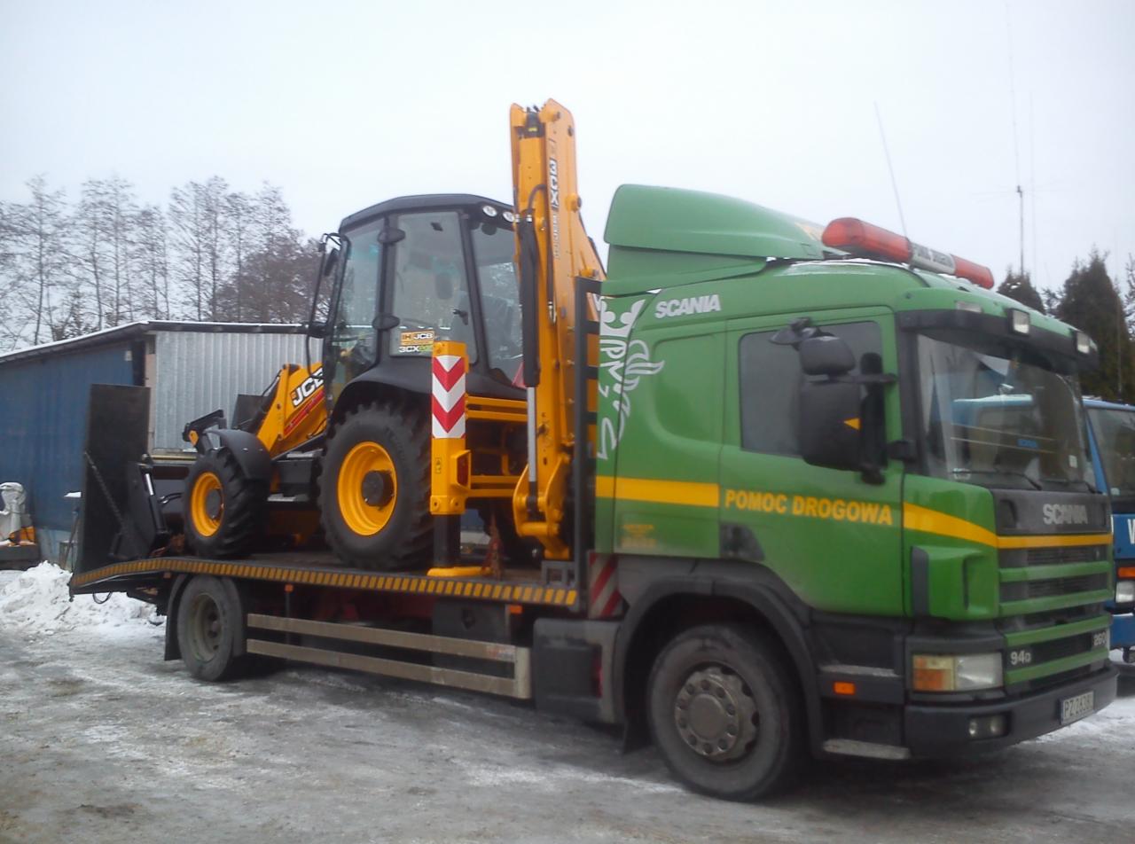 Zielona laweta Scania z żółtą koparko-ładowarką JCB na platformie. Widoczny napis 'Pomoc Drogowa' i sygnały świetlne. Zimowy krajobraz w tle.