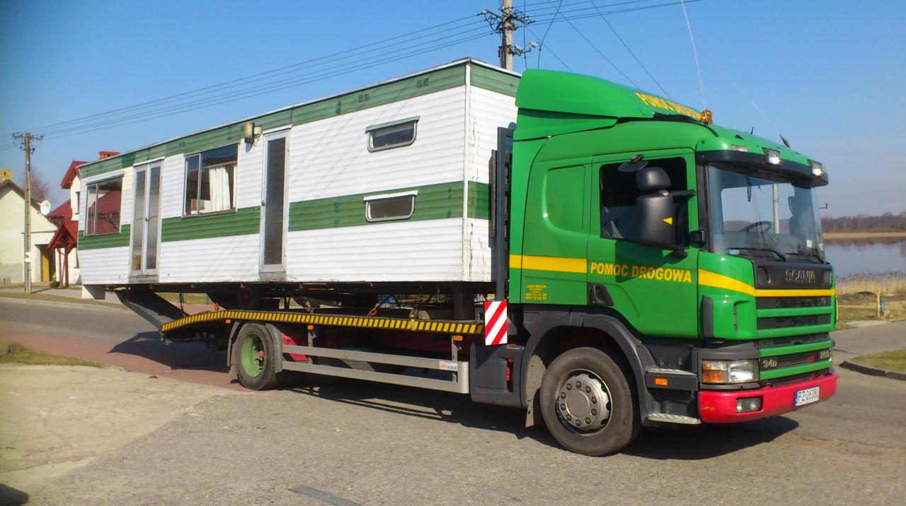 Zielona laweta Scania z napisem 'Pomoc Drogowa' przewozi biało-zielony domek mobilny na niskopodwoziowej platformie. Widok z boku, słoneczny dzień.