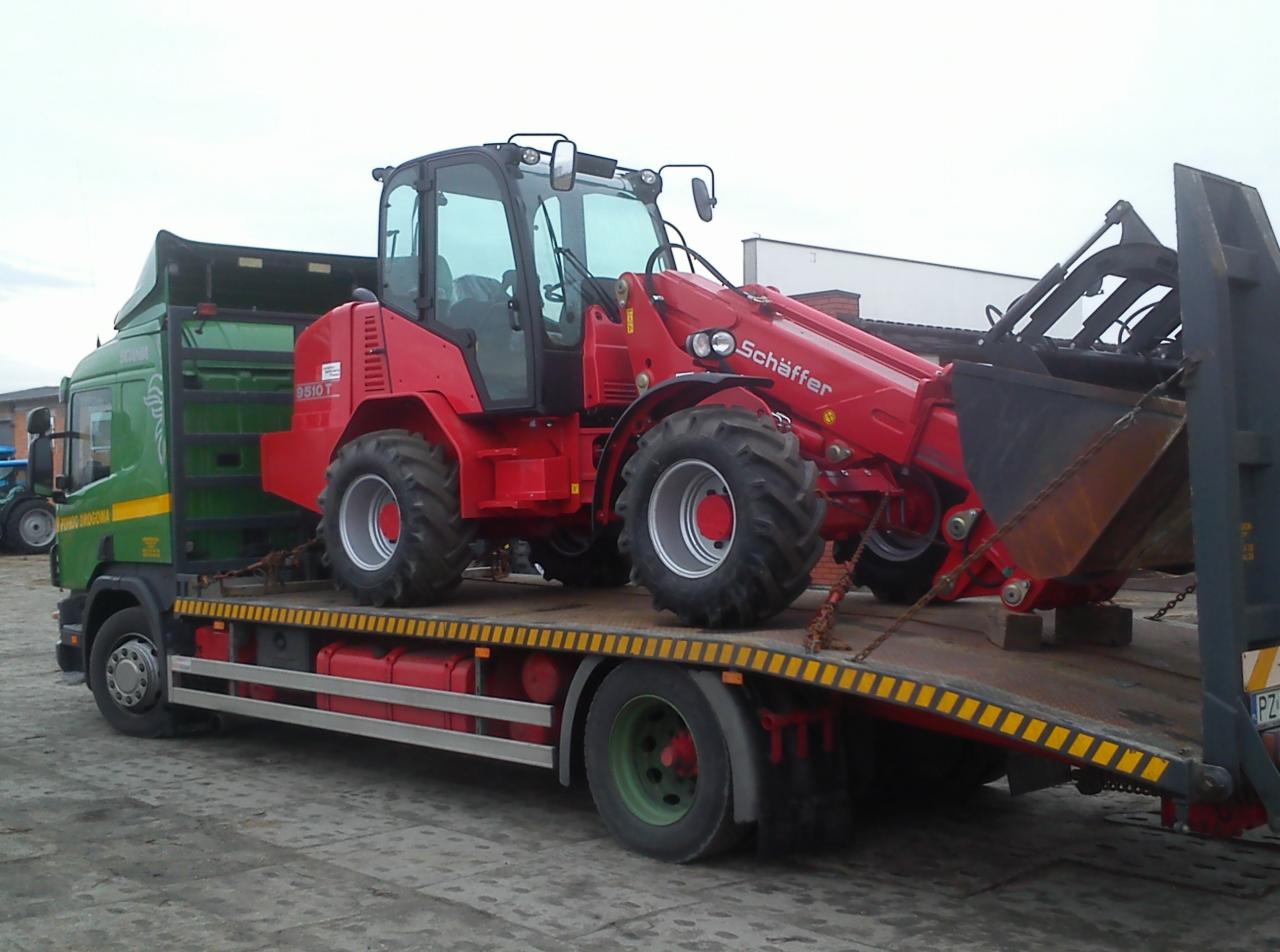 Zielona ciężarówka z czerwoną ładowarką teleskopową Schäffer 9510T na platformie. Ładunek zabezpieczony łańcuchami, widoczne detale opon i konstrukcji pojazdów.