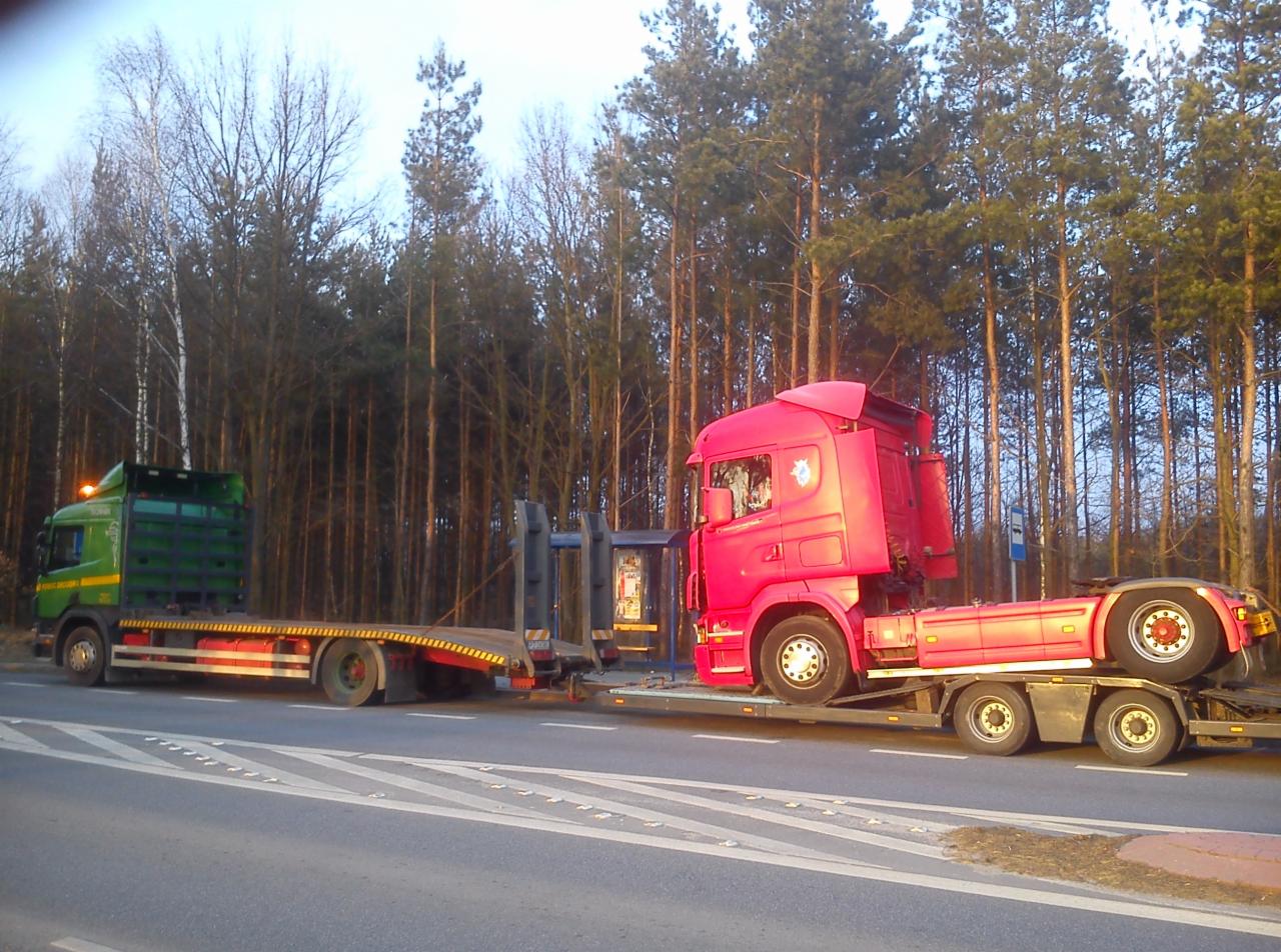 Dwa samochody ciężarowe typu laweta: zielony i czerwony, jadące obok siebie po drodze. W tle wysokie drzewa. Czerwony pojazd przewozi kabinę ciężarówki.