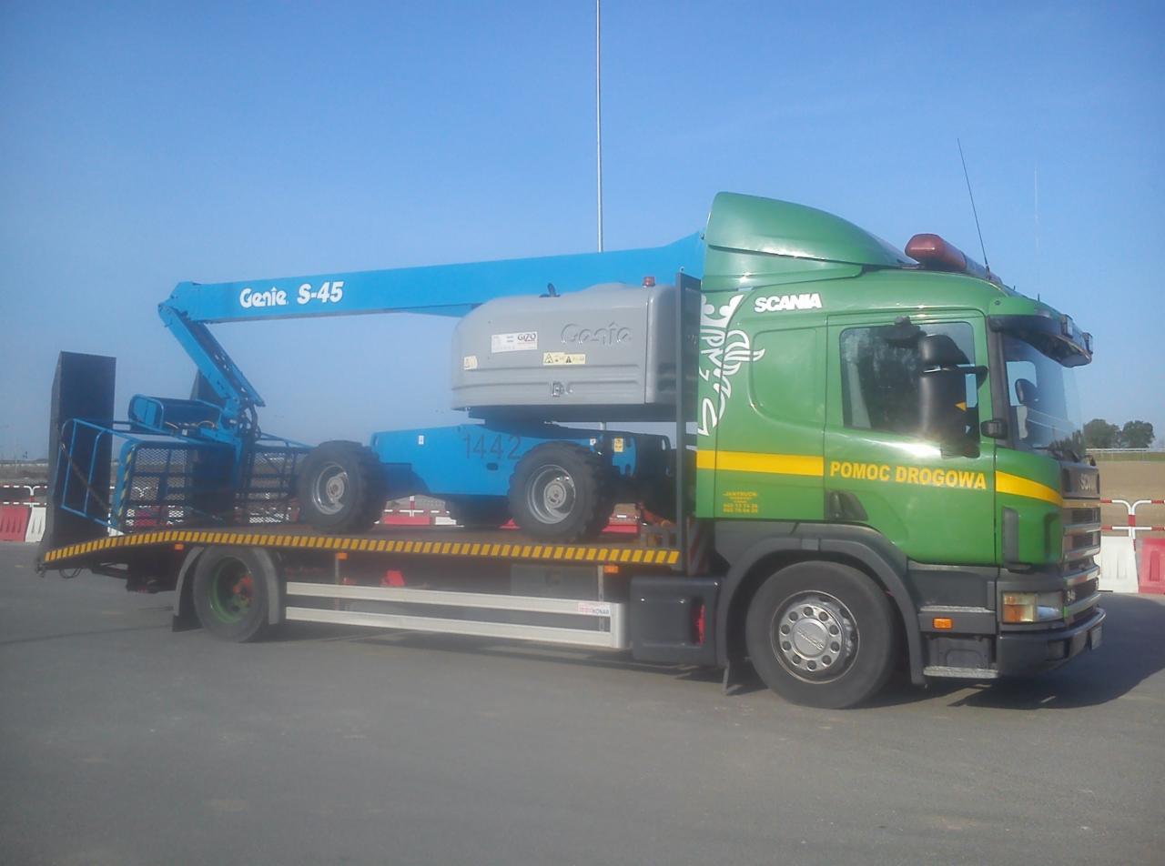 Laweta z podnośnikiem Genie S-45 na platformie. Zielona kabina Scania z napisem 'Pomoc Drogowa'. Błękitne niebo w tle. Widok z boku, światło dzienne.