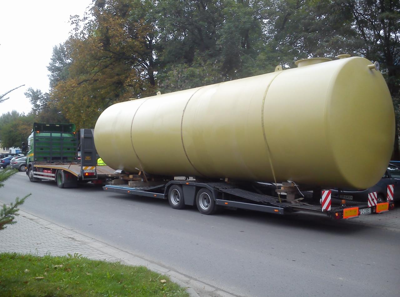 Żółty zbiornik przewożony na niskopodwoziowej lawecie, widok z boku na ulicy. Tylne światła lawety i zielona kabina ciężarówki widoczne w tle.