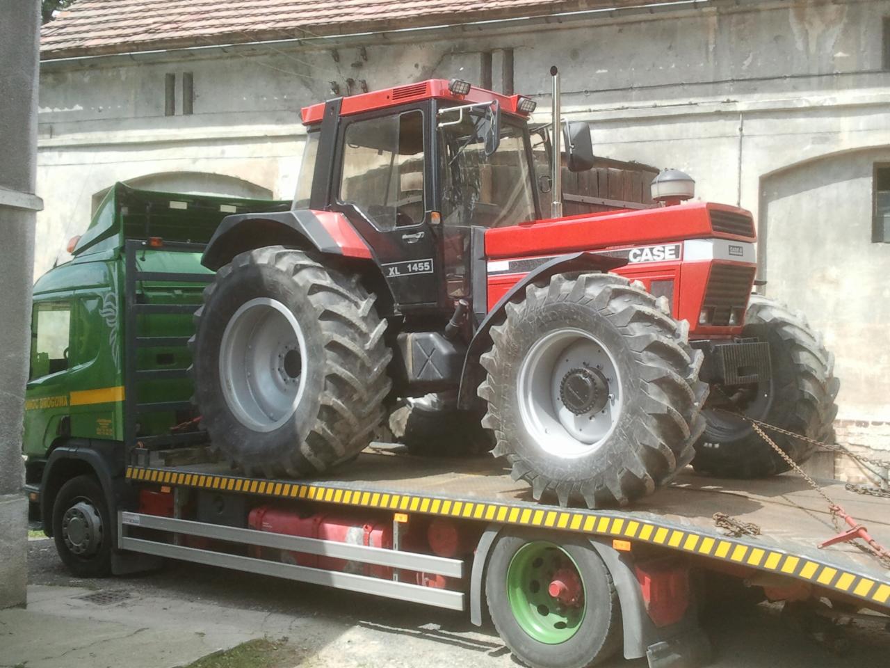 Czerwony ciągnik Case XL 1455 na zielonej lawecie z żółtymi pasami, transportowany w dzień. Widoczne budynki w tle.