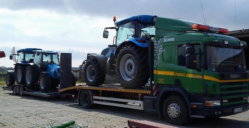 Zielona laweta Scania z napisem 'Pomoc Drogowa' przewozi dwa niebieskie traktory. Jeden traktor na lawecie, drugi na przyczepie. Słoneczny dzień.