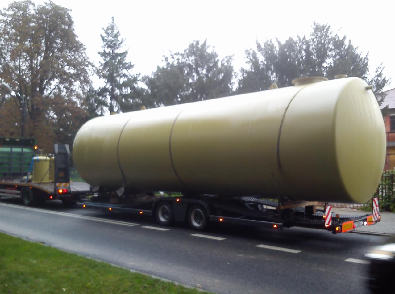 Żółty, cylindryczny zbiornik na niskopodwoziowej platformie transportowej, przewożony po asfaltowej drodze, w tle drzewa i budynek.