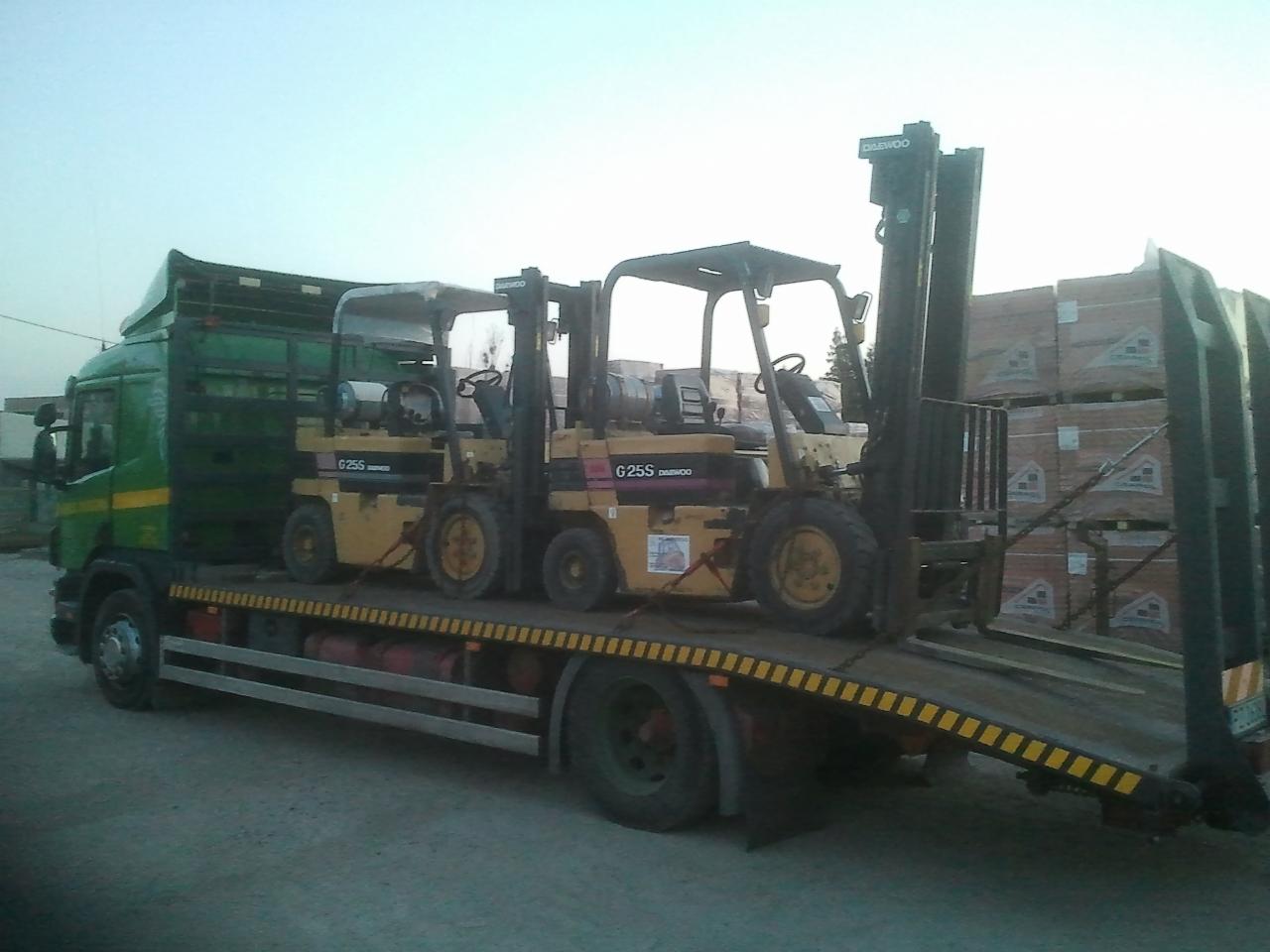 Zielono-żółta ciężarówka przewozi dwa wózki widłowe Daewoo G25S na platformie. W tle palety z materiałami budowlanymi. Transport maszyn budowlanych.