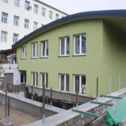 Budynek w trakcie budowy z widocznym fundamentem i przygotowaniem do instalacji, zielona elewacja, białe okna.