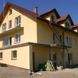 Dwupiętrowy, otynkowany na żółto dom z brązowym dachem i lukarnami, widoczny z zewnątrz, z elementami rusztowania oraz stertą cegieł przed budynkiem, na tle błękitnego nieba.