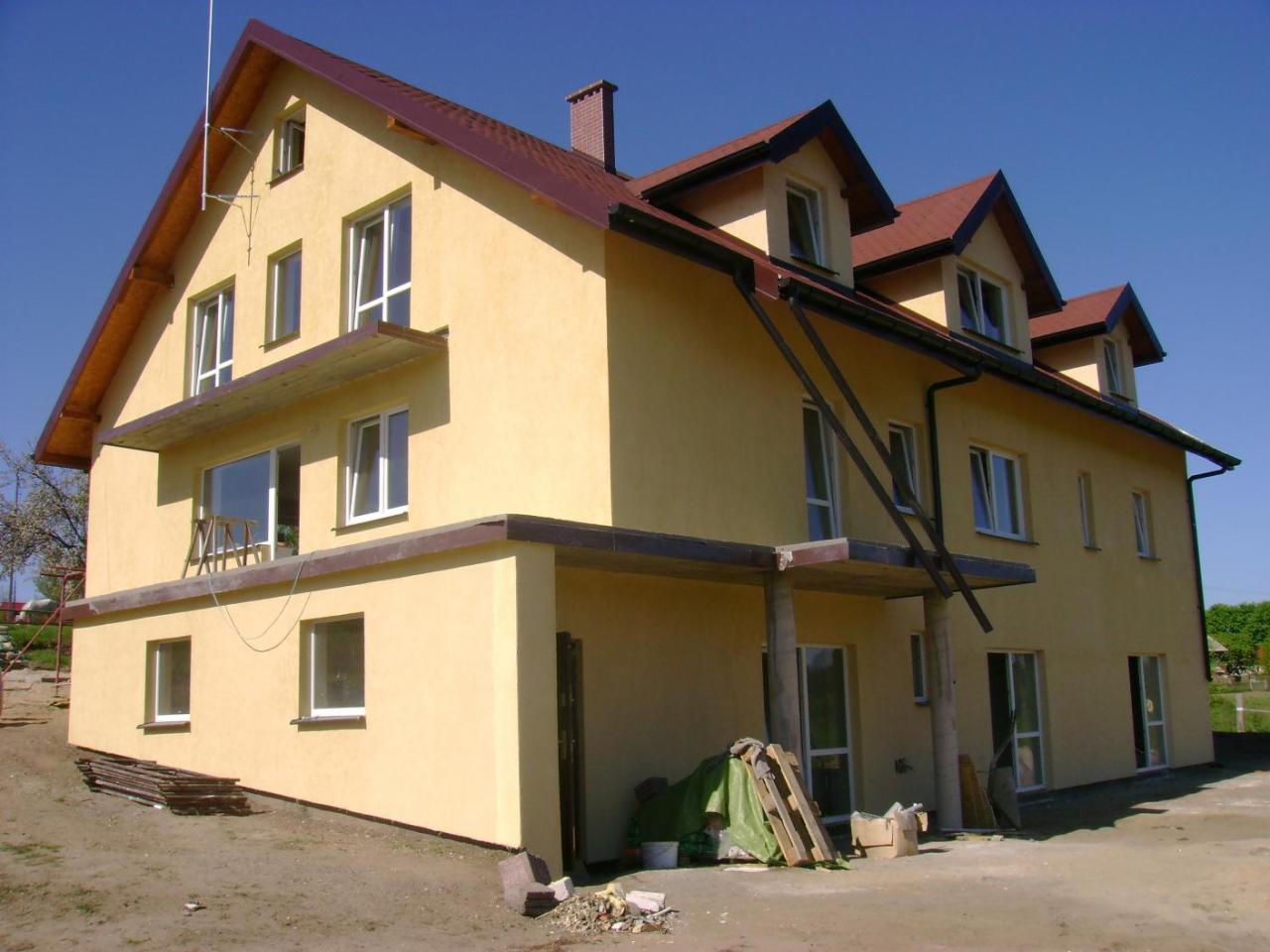 Dwupiętrowy, otynkowany na żółto dom z brązowym dachem i lukarnami, widoczny z zewnątrz, z elementami rusztowania oraz stertą cegieł przed budynkiem, na tle błękitnego nieba.