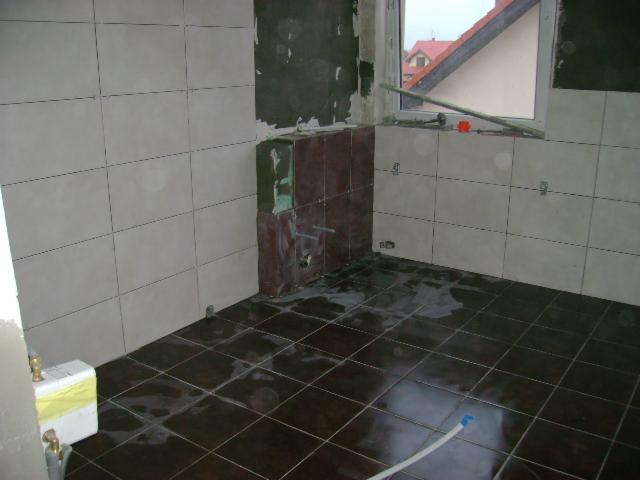 Wnętrze łazienki w trakcie prac wykończeniowych: częściowo wyłożone jasnymi płytkami ściany, ciemna, mokra podłoga z płytek, widoczne elementy instalacji i okno z widokiem na dach budynku.