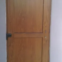 Brązowe drzwi zewnętrzne z czarnymi zawiasami, widoczne ślady zaprawy murarskiej na podłodze i ścianie.